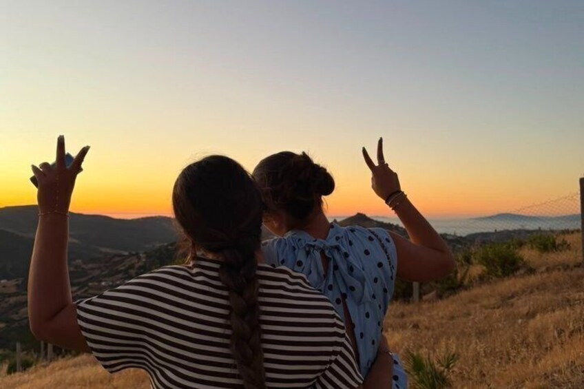 Two people with their backs to the camera, making peace signs, overlooking a sunset with a mountainous landscape in the background. 2