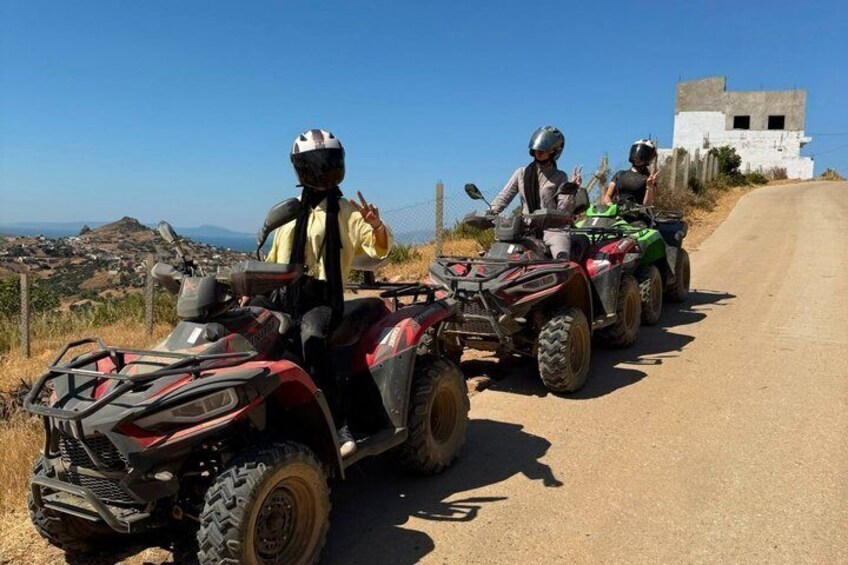 Two people on ATVs on a dirt road with a white building in the background. 1