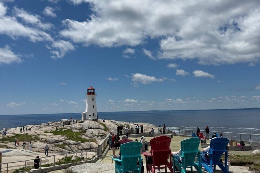 A lighthouse on a rocky outcrop with people walking and sitting on chairs by the water. 2