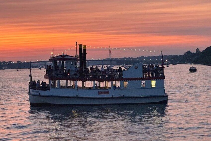 A boat with people on deck, a sunset in the background, and a bridge in the distance. 1