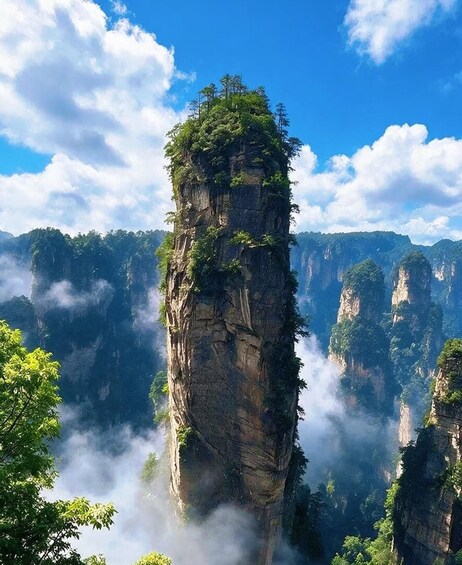 A tall, narrow rock formation with dense vegetation on top, surrounded by misty mountains. 1