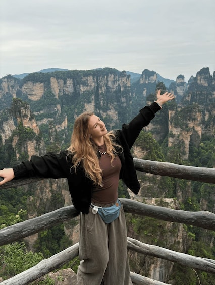 A person standing with arms outstretched, overlooking a mountainous landscape with steep cliffs and dense greenery. 1