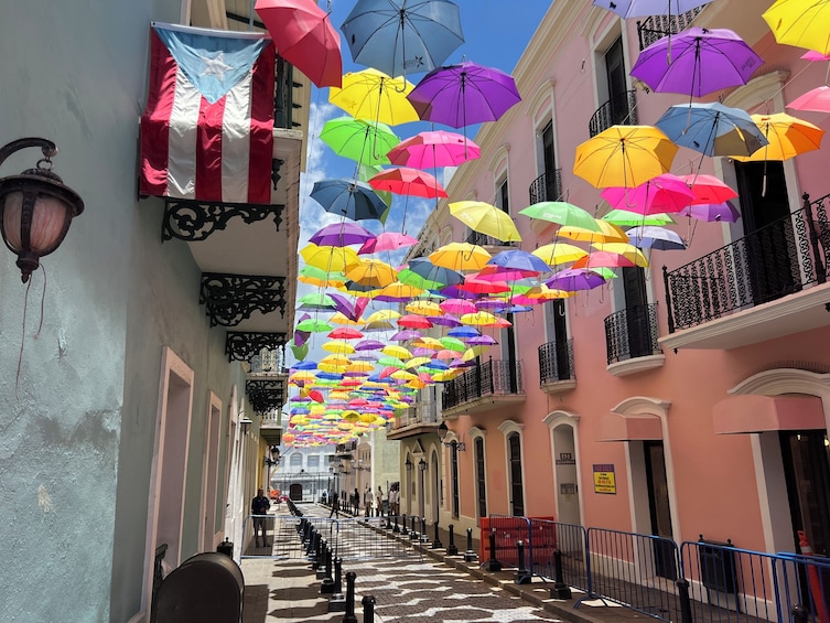 A street with colorful umbrellas hanging overhead, buildings with balconies, and a clear blue sky. 1