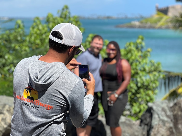 A person taking a photo of a couple by a coastal area with greenery and a clear view of the sea. 2