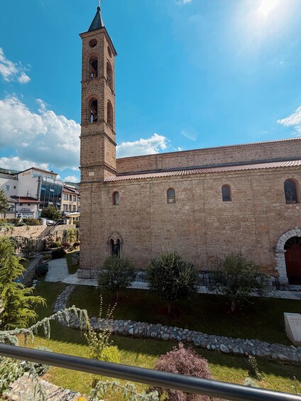 A historic stone building with a bell tower, surrounded by a well-maintained garden and a clear blue sky. 2