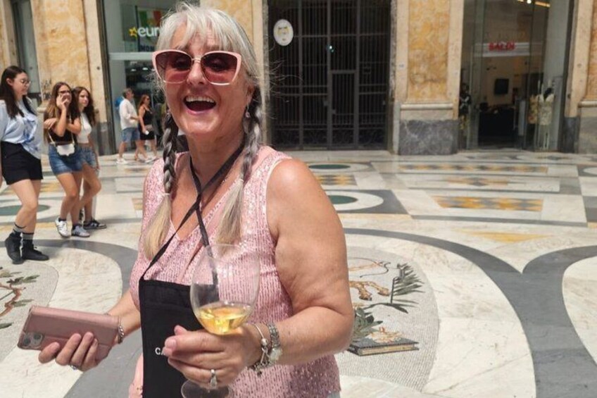 A person holding a glass of wine and a phone, standing in a public square with patterned pavement and buildings in the background. 1