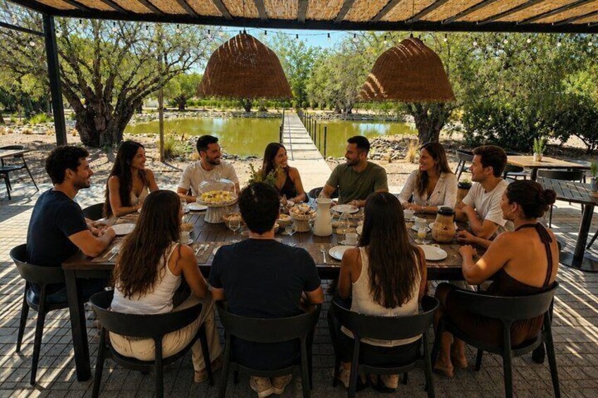 A group of people dining at an outdoor table with a thatched roof, surrounded by trees and a body of water. 4