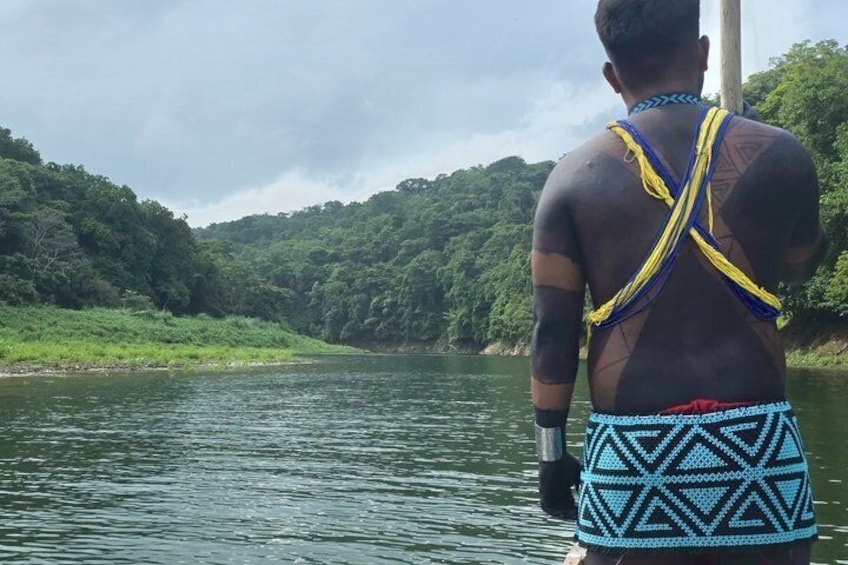 A person standing on a boat, wearing a patterned garment, with a river and forested hills in the background. 1