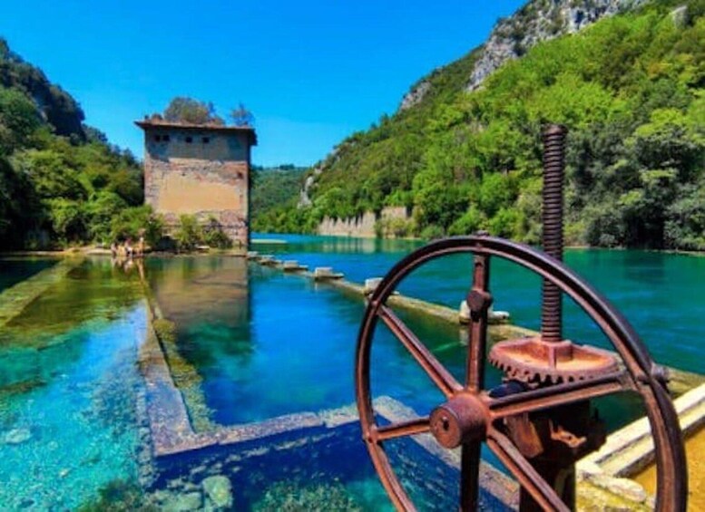 A clear blue lake with a rustic water wheel, a stone tower, and lush green hills. 2