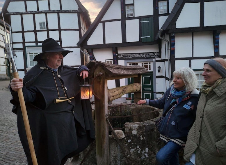 A person in a black cloak holding a lantern and a stick, standing in front of a traditional half-timbered building. 1