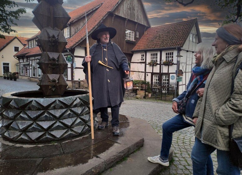 A historical reenactment scene with a person in period costume holding a staff and a lantern, standing in front of a traditional building with a cobblestone street. 4