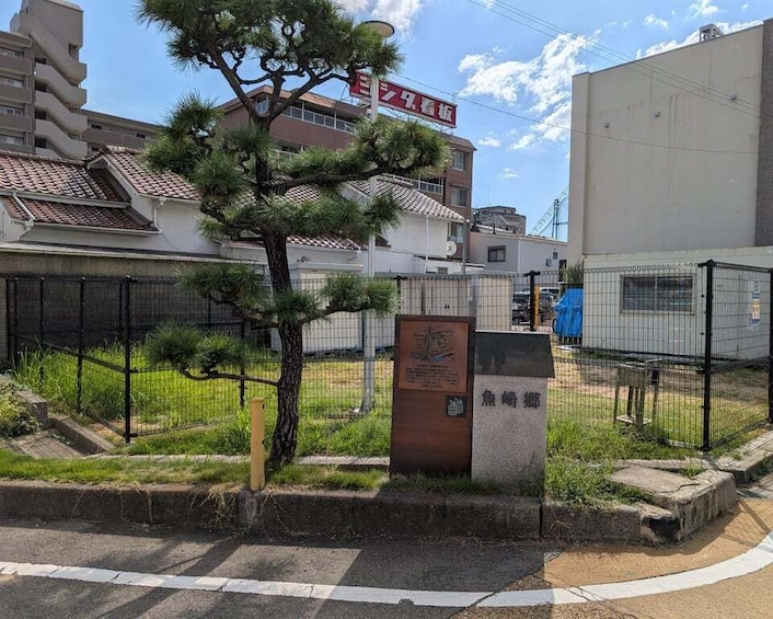 A fenced area with a tree, a stone monument, and a building with a red sign in the background. 3