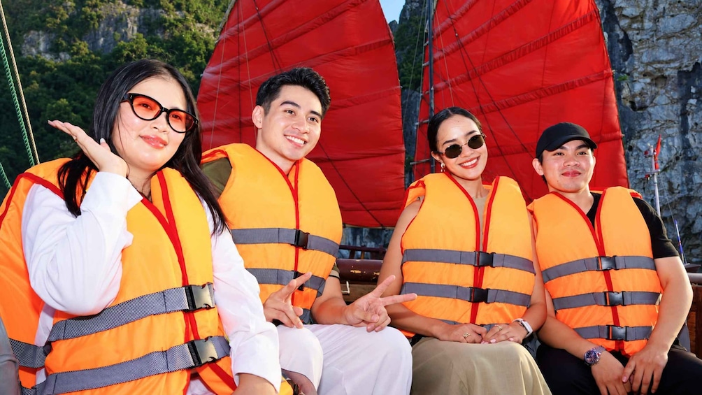 Four people wearing orange life jackets, sitting on a boat with red sails. 2