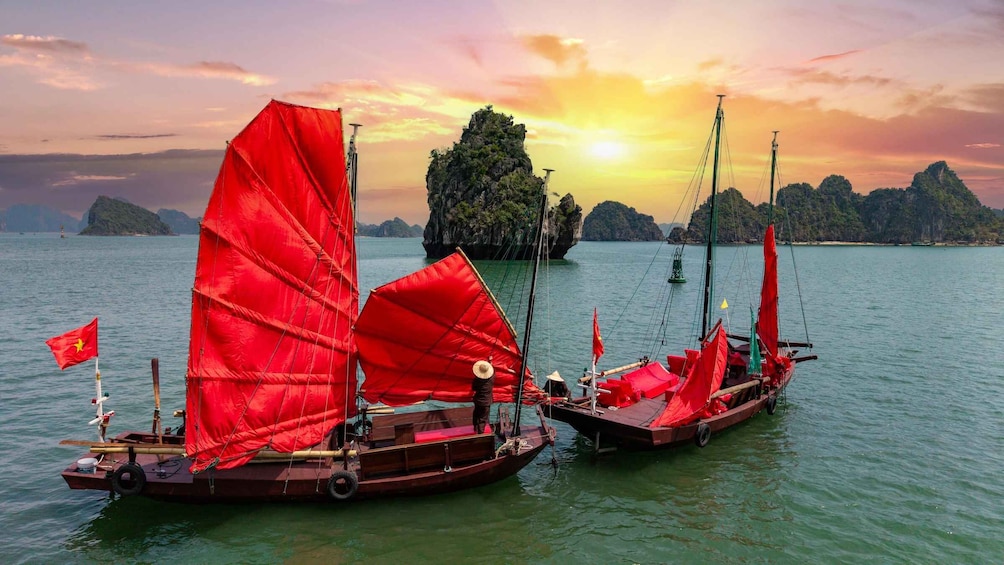 Two traditional boats with red sails on a calm sea with rocky islands in the background during sunset. 1