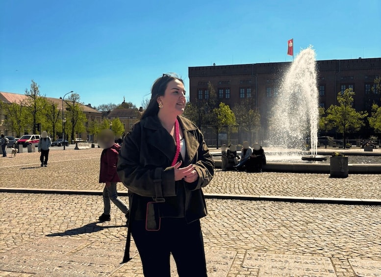 A woman in a jacket and scarf standing in a public square with a fountain and buildings in the background. 4
