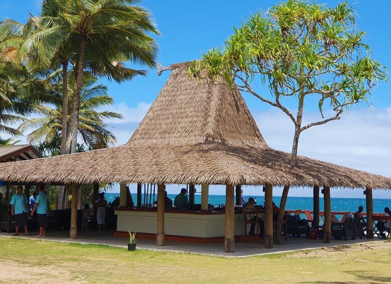 A thatched-roof pavilion with people sitting and standing around it, overlooking a beach with palm trees and the ocean. 3
