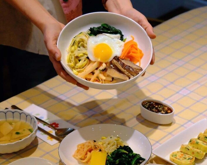 A person holding a bowl of food with various ingredients like noodles, egg, vegetables, and meat. 1