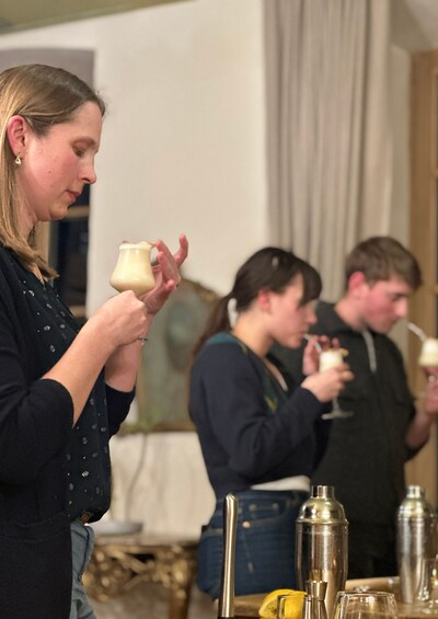 A person holding a small container, possibly tasting a beverage, with a table of various bottles and glasses in the background. 3