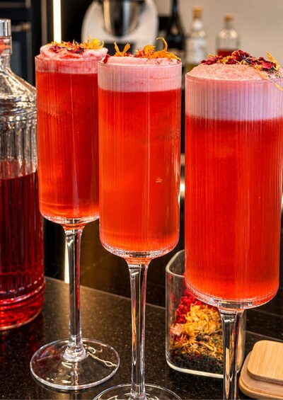Three tall glasses of a red beverage, possibly a cocktail, garnished with orange zest and flowers. 1