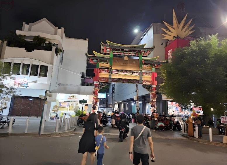 A street at night with a traditional Asian gate, shops, and people walking. 1