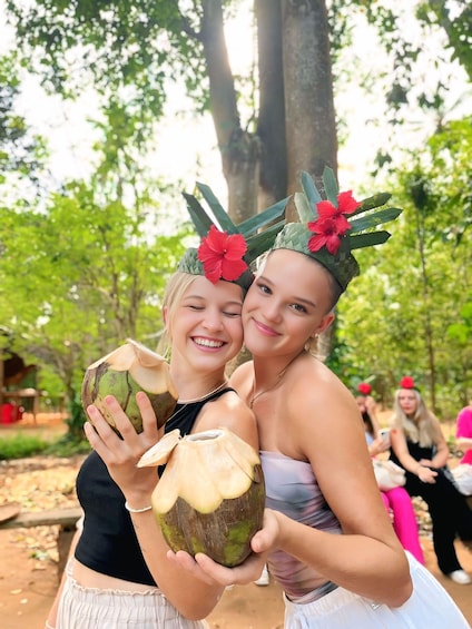 Two people holding coconuts with carved tops, wearing floral headpieces, in a forested outdoor setting. 1