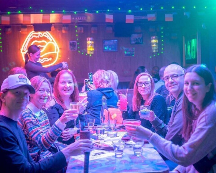 A group of people at a neon-lit bar with drinks and a neon sign in the background. 1