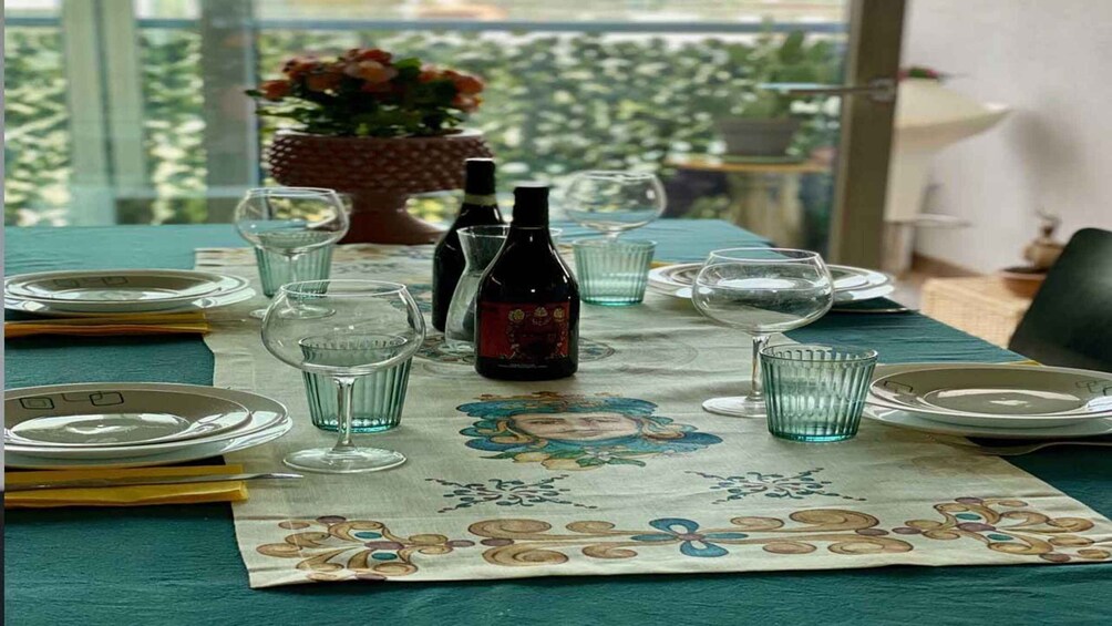 A table set for a meal with plates, glasses, and a bottle of wine, with a floral tablecloth and a view of greenery outside. 4