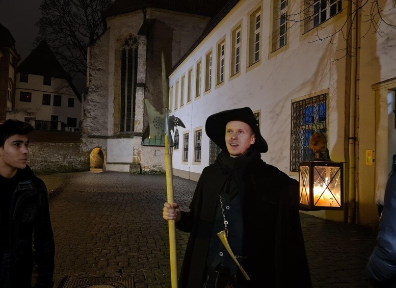 A person in historical costume holding a staff and a trumpet, standing in a cobblestone street with old buildings and a church in the background. 4