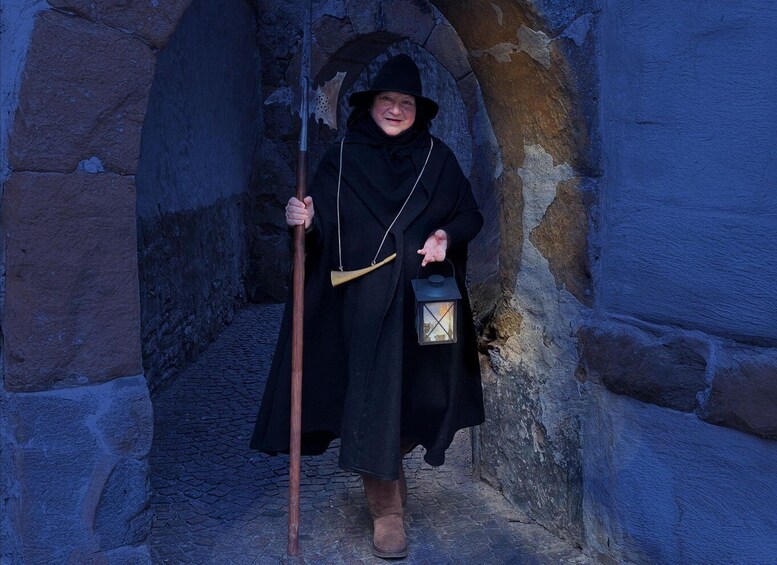 A person in a long black coat holding a walking stick and a lantern, standing in a stone archway. 3
