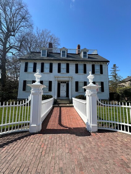 A white colonial-style house with a brick pathway leading to the front door, flanked by white picket fences. 1