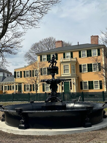 A black fountain with a statue in a park, surrounded by a green fence, with a yellow building in the background. 2