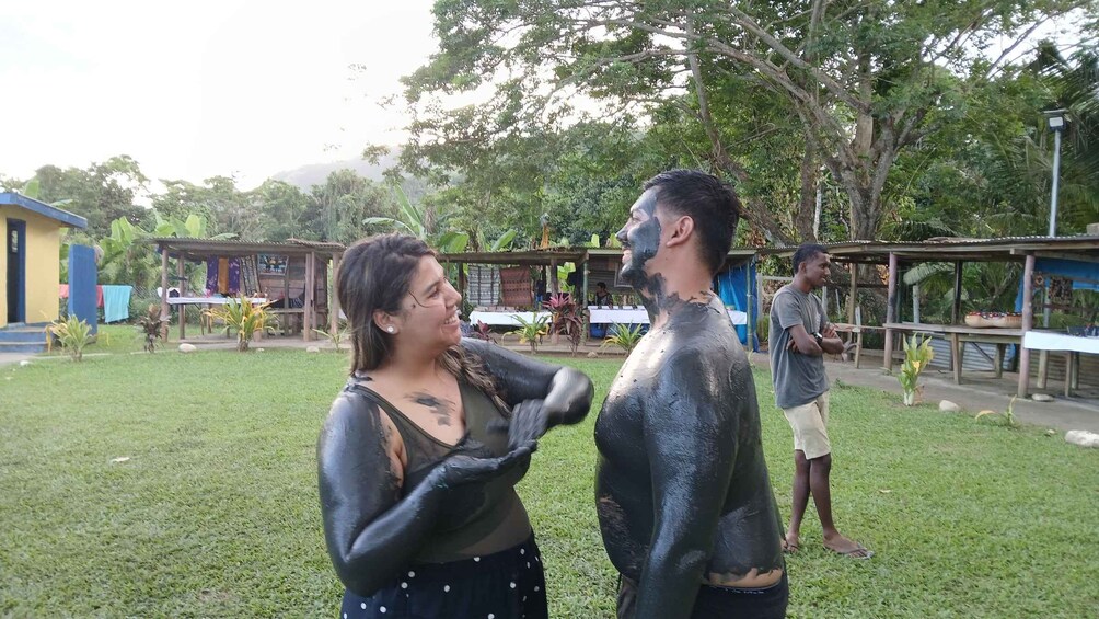 Two people covered in black substance, standing on grass with buildings and trees in the background. 3