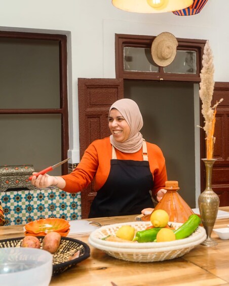 A person in an orange top and black apron preparing food at a kitchen counter with various vegetables and a large clay pot. 5