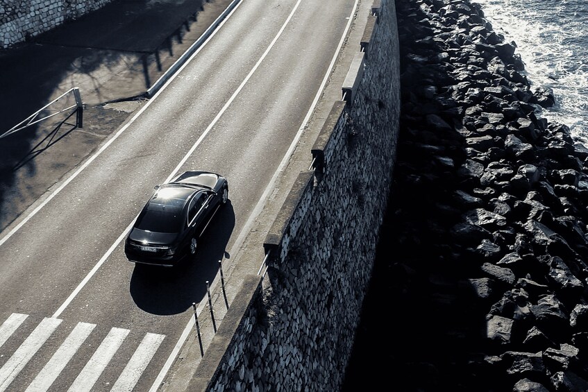 A black car driving on a road with a stone wall on one side and a rocky shoreline on the other. 3