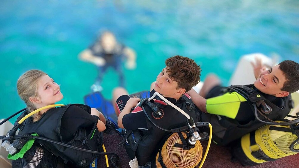 Three individuals in scuba diving gear sitting on a boat, with a diver in the background. 2