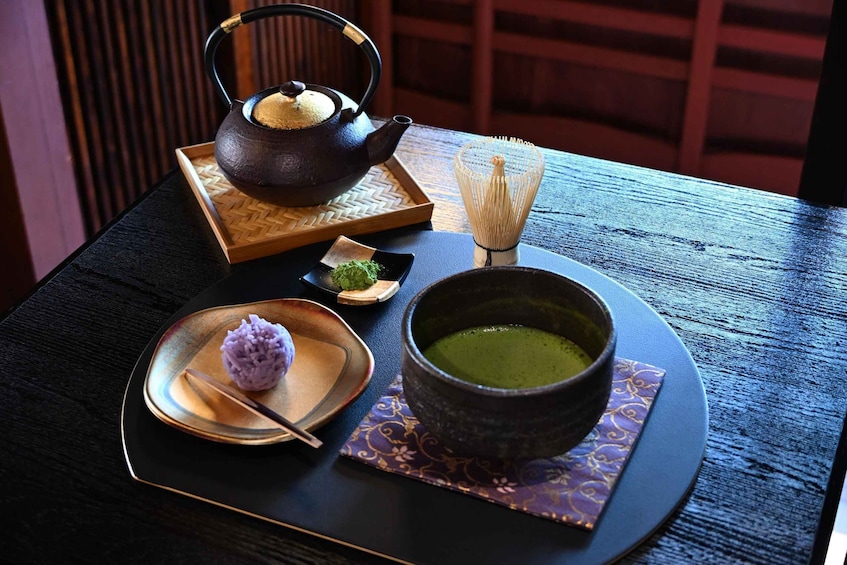 A traditional Japanese tea ceremony setup with a tea pot, a bowl of green tea, a small dish with a purple dessert, and a whisk. 5
