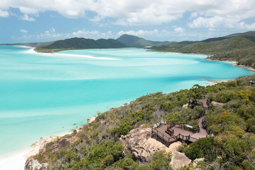 A coastal landscape with turquoise waters, a sandy beach, and a forested hill with a wooden viewing platform. 3