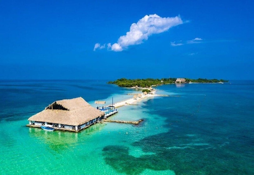 An overwater bungalow with a thatched roof, clear turquoise waters, and a small island with a building in the distance. 4