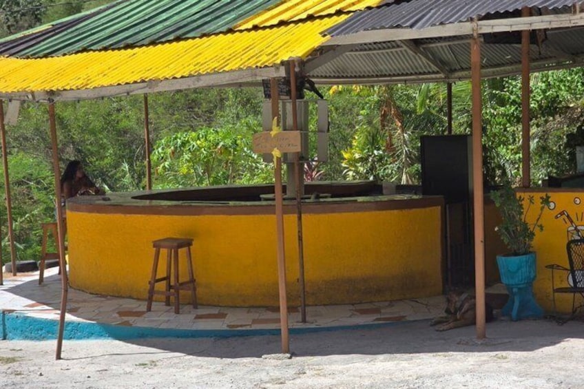 A yellow outdoor bar with a metal roof, a wooden stool, and a potted plant. 5