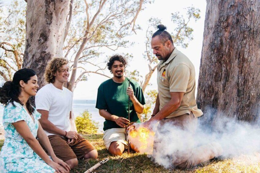 A group of people around a campfire outdoors with trees and a body of water in the background. 1