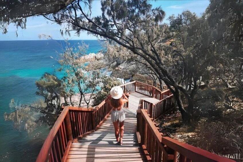 A wooden boardwalk leading to a beach with clear turquoise water, surrounded by trees and rocky outcrops. 3