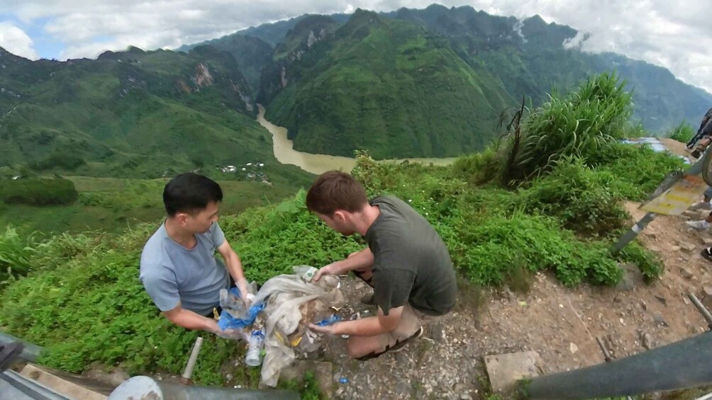 Zwei Personen betrachten eine Karte und eine Plastiktüte auf einem Bergpfad. Im Hintergrund fließt ein Fluss durch üppiges Grün. 1