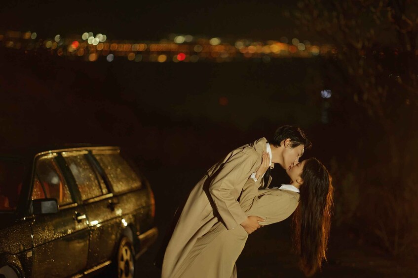 A couple kissing in front of a car at night with a bridge in the background. 3