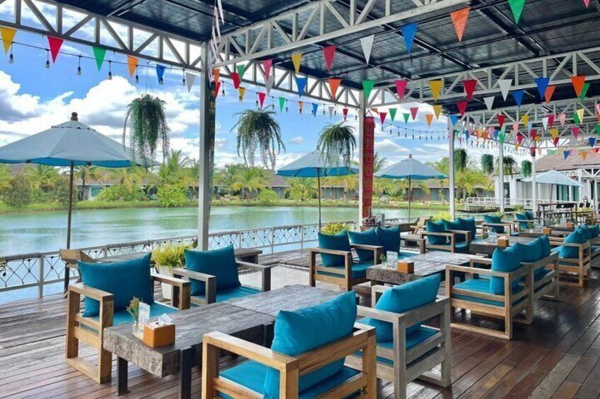 A covered outdoor seating area with wooden furniture and blue cushions, overlooking a body of water with palm trees and umbrellas. 3