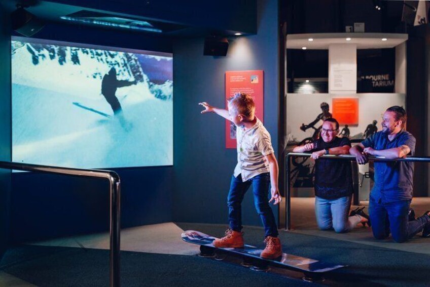 A person snowboarding indoors on a platform with a large screen displaying a snowboarder in motion. 1