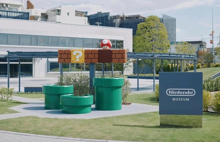 A Nintendo Museum entrance with a Mario statue, green planters, and modern buildings in the background. 1