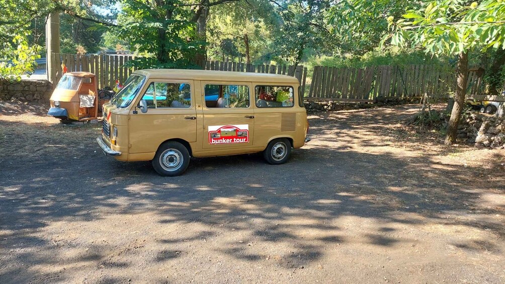 A vintage van parked on a dirt path with a wooden fence and trees in the background. 1
