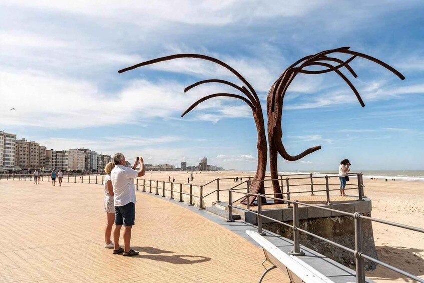 A beach with a large metal sculpture, people walking, and buildings in the background. 2