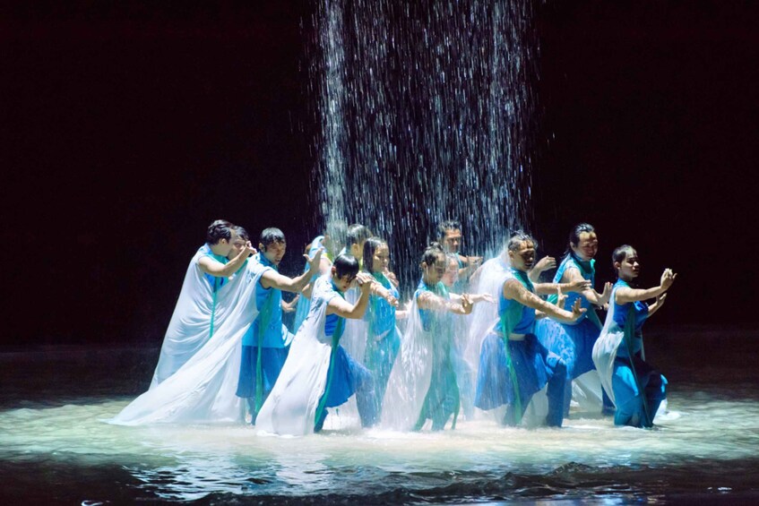 A group of people in blue and white costumes performing a synchronized dance on a stage with a water fountain in the background. 1