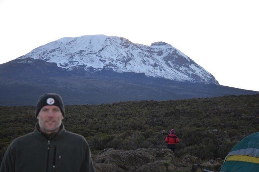 A person in a dark jacket and beanie, a red jacket person in the distance, and a snow-capped mountain. 2